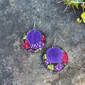May include: A pair of fabric earrings with a grape design. Each earring features a circular base with a black background and a pattern of purple grapes, red berries, and green leaves. A smaller, solid purple circle is sewn on top.