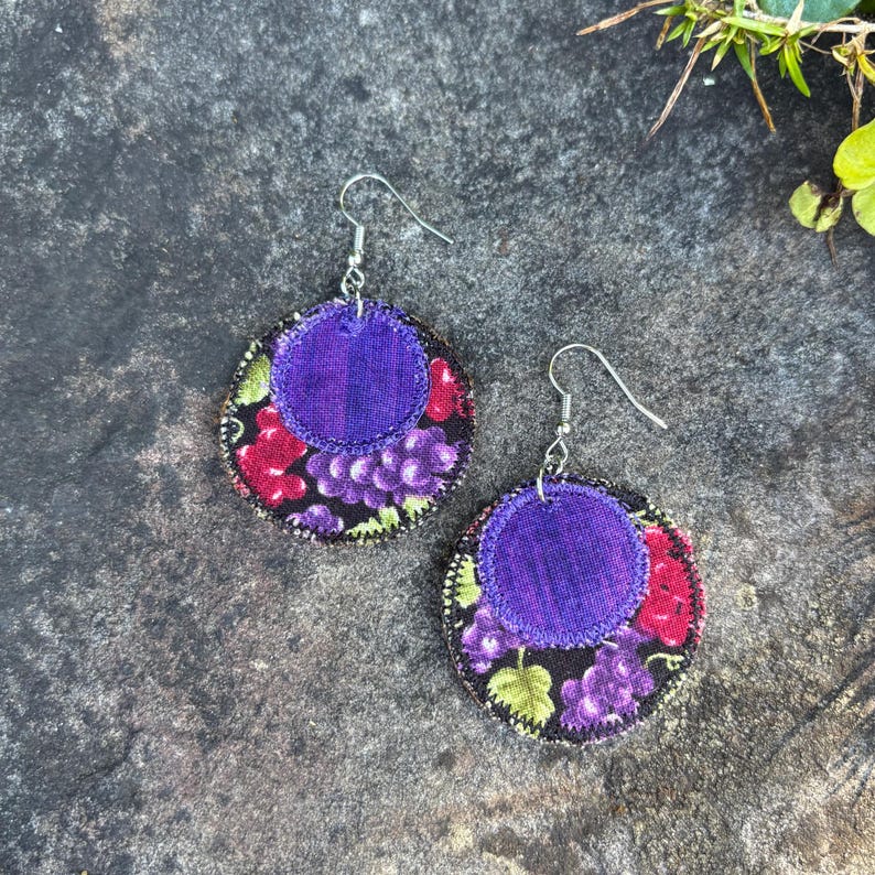 May include: A pair of fabric earrings with a grape design. Each earring features a circular base with a black background and a pattern of purple grapes, red berries, and green leaves. A smaller, solid purple circle is sewn on top.