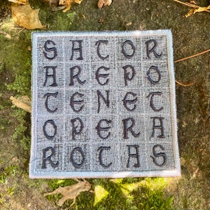 May include: A square, gray fabric patch with embroidered black letters arranged in a grid. The text reads "SATOR AREPO TENET OPERA ROTAS". The patch has a light blue border and is set against a natural, mossy background.