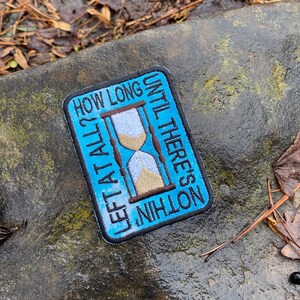 May include: Embroidered patch with a blue background and the text "HOW LONG LEFT AT ALL? UNTIL THERE'S NOTHING". The patch features a brown and beige hourglass design. The patch is rectangular with a black border.