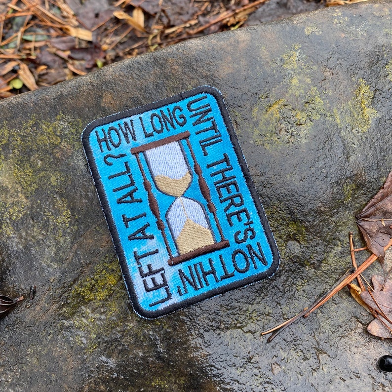 May include: Embroidered patch with a blue background and the text "HOW LONG LEFT AT ALL? UNTIL THERE'S NOTHING". The patch features a brown and beige hourglass design. The patch is rectangular with a black border.