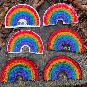 May include: Six embroidered rainbow patches with a white border and the text "BMFS" on some. The rainbows feature vibrant colors: red, orange, yellow, green, blue, and purple. The patches are arranged on a stone surface.