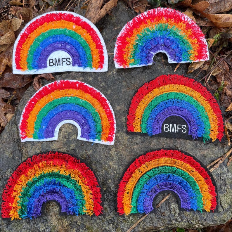 May include: Six embroidered rainbow patches with a white border and the text "BMFS" on some. The rainbows feature vibrant colors: red, orange, yellow, green, blue, and purple. The patches are arranged on a stone surface.