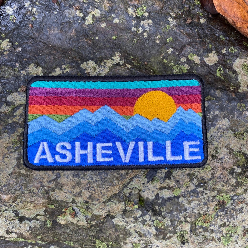 Peut inclure: &Eacute;cusson brod&eacute; repr&eacute;sentant un paysage de montagne avec un coucher de soleil et le mot "ASHEVILLE" en blanc. Le patch a une bordure noire et un design color&eacute; avec des nuances de bleu, orange, jaune et violet.