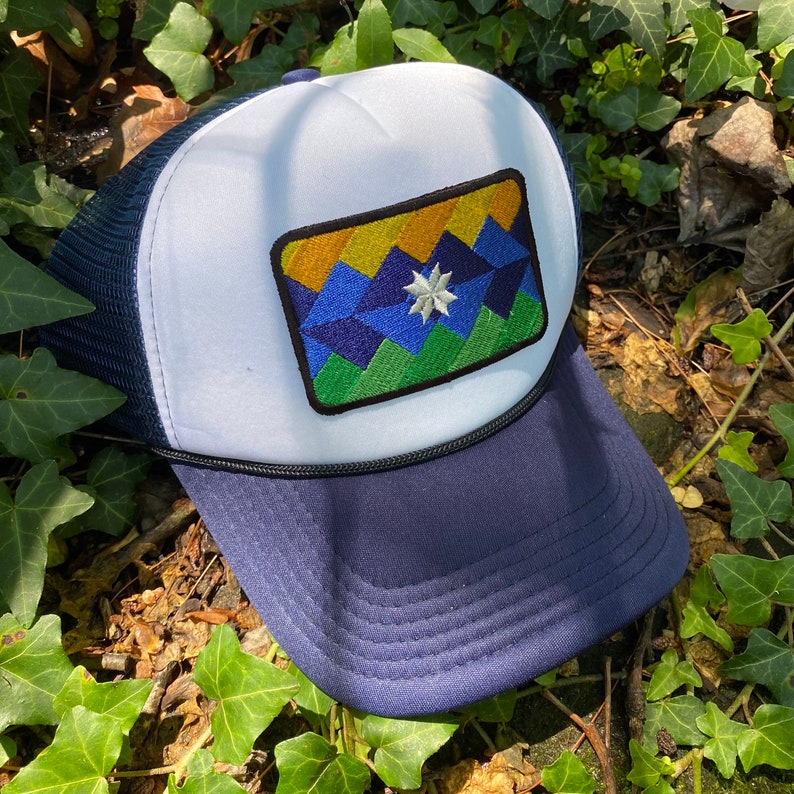 May include: A navy blue and white trucker hat with a geometric patch featuring a mountain design in shades of blue, green, yellow, and a white star. The hat has a mesh back and a curved brim.