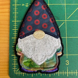 May include: A fabric gnome patch with a navy blue hat featuring red circles, a gray beard, and a green and purple outfit. The patch has a black border and is on a green cutting mat.