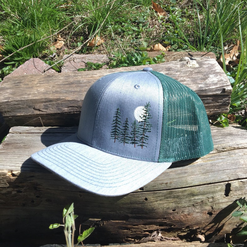 May include: A gray and green trucker hat with an embroidered forest and moon design. The front panel is gray with a forest and moon design, and the back is green mesh. The brim is gray.