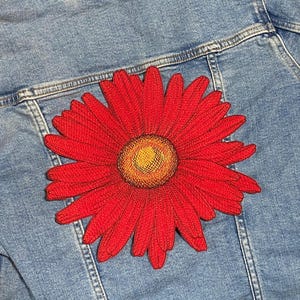 May include: A large, embroidered red daisy patch with a yellow center is affixed to the back of a light blue denim jacket. The patch is centered on the jacket and adds a pop of color.