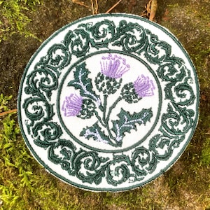 May include: Embroidered round patch featuring a thistle design. The patch has a white background with a green border and a central image of purple thistles with green leaves. The border has a decorative scroll pattern.
