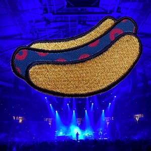 May include: Embroidered patch featuring two hot dogs. The hot dogs have a golden-yellow bun and a blue top with red circular designs. The background is a concert venue with blue lighting.