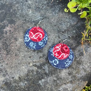 May include: A pair of handmade fabric earrings. Each earring features a red paisley center, surrounded by a navy blue floral pattern. The earrings have silver-colored ear wires and are displayed on a gray stone surface.
