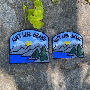 May include: Two embroidered patches with the phrase "Ain't Life Grand" above a mountain and lake scene. The patches feature a blue sky, gray mountains, green trees, and a yellow sun. The patches are on a gray stone surface.
