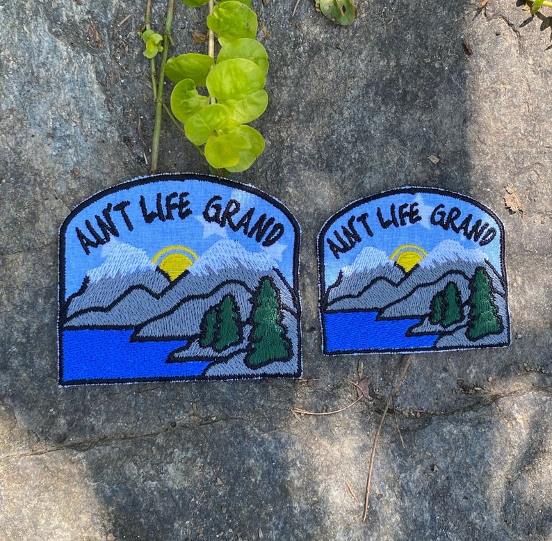 May include: Two embroidered patches with the text "Ain't Life Grand" above a mountain and lake scene. The patches feature a blue sky, grey mountains, green trees, and a yellow sun. The patches are on a grey stone surface.