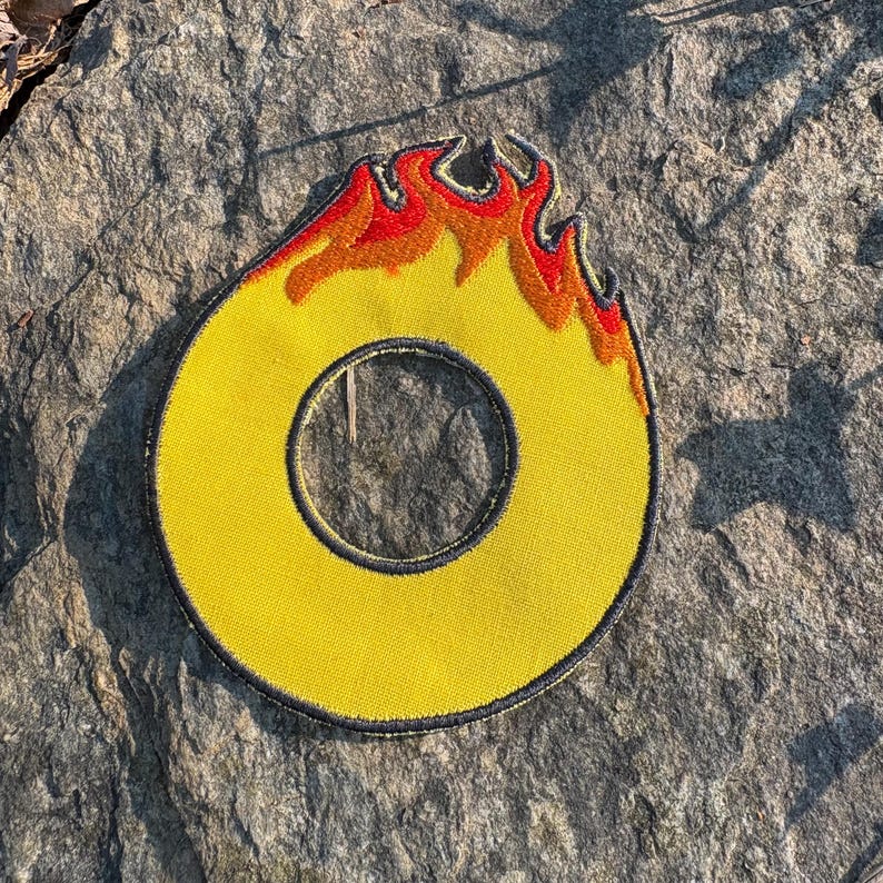 Puede incluir: Parche bordado amarillo con forma de la letra "O" con llamas. Las llamas son rojas y naranjas, y el parche tiene un borde negro. El parche es circular con un agujero en el centro.