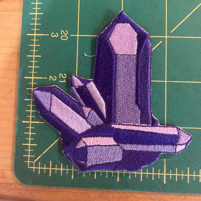 May include: Embroidered patch depicting a cluster of crystals in shades of purple and lavender. The patch has a detailed design with various facets and textures, suitable for adding to clothing or accessories.
