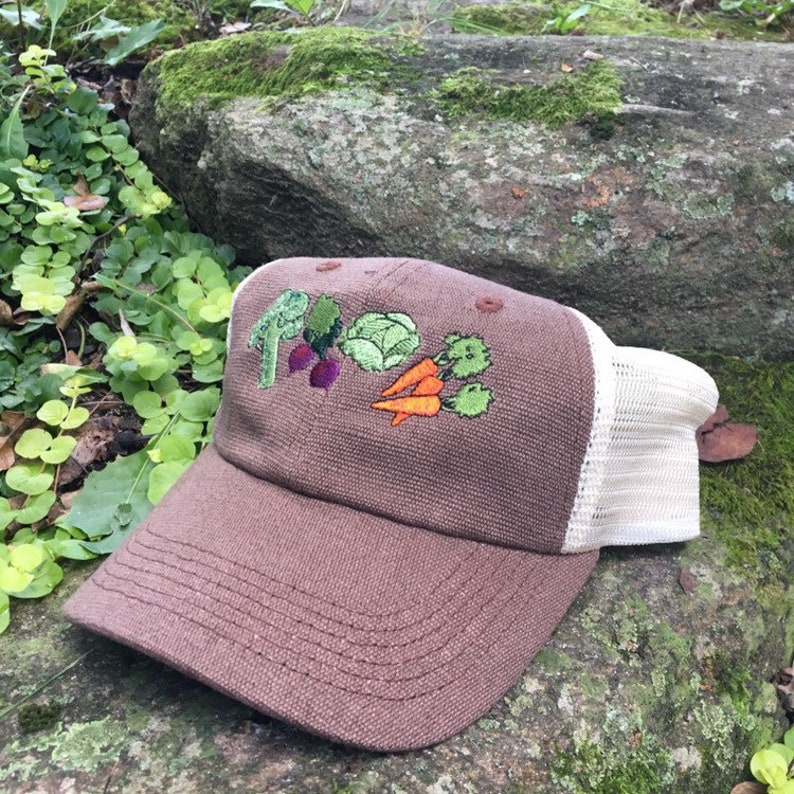 May include: Brown baseball cap with a white mesh back. The front of the cap features embroidered vegetables: broccoli, beets, cabbage, carrots, and cauliflower. The cap is sitting on a mossy rock.
