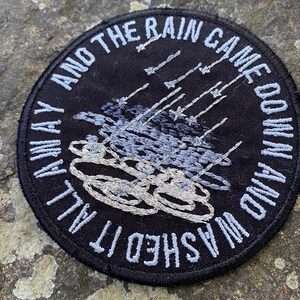 Pode incluir: Um patch redondo bordado em preto com texto branco: "AND THE RAIN CAME DOWN AND WASHED IT ALL AWAY." O design apresenta chuva e nuvens bordadas em tons de cinza e branco. Ideal para jaquetas ou bolsas.