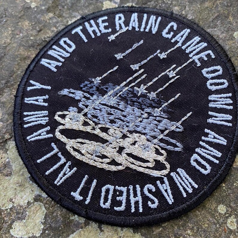 Pode incluir: Um patch redondo bordado em preto com texto branco: "AND THE RAIN CAME DOWN AND WASHED IT ALL AWAY." O design apresenta chuva e nuvens bordadas em tons de cinza e branco. Ideal para jaquetas ou bolsas.