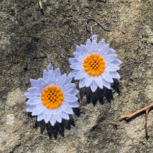 May include: A pair of daisy-shaped earrings. Each earring features a white, lace-like flower with a bright yellow center. The earrings have silver-colored hooks for pierced ears. The earrings are set against a textured, gray stone surface.