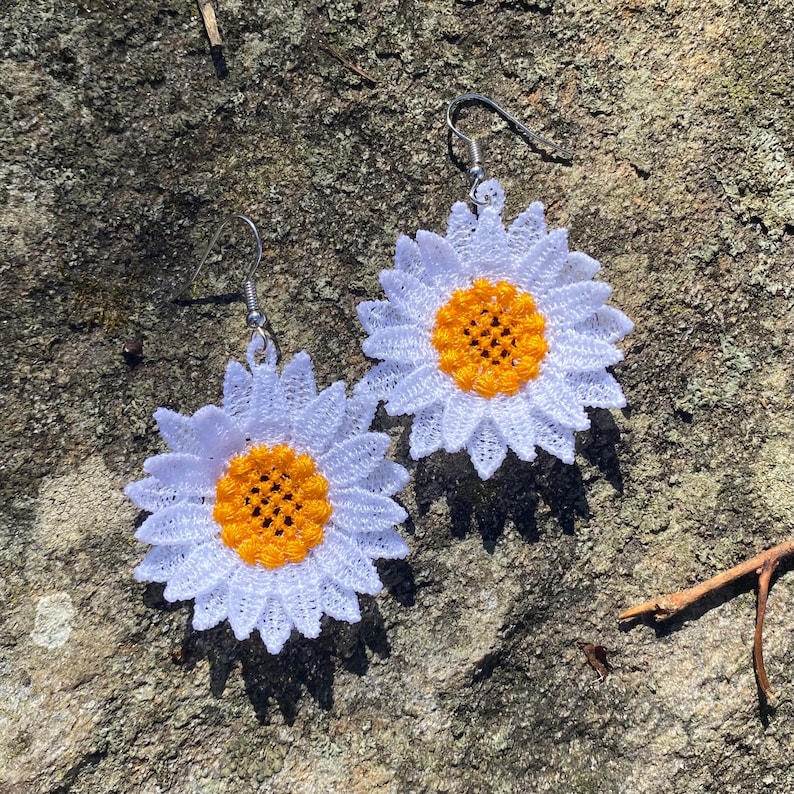May include: A pair of daisy-shaped earrings. Each earring features a white, lace-like flower with a bright yellow center. The earrings have silver-colored hooks for pierced ears. The earrings are set against a textured, gray stone surface.