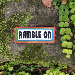 May include: A rectangular patch with the words "RAMBLE ON" in black block letters. The patch has a white background with a blue, green, and red border. The patch is set on a stone surface surrounded by green plants and moss.