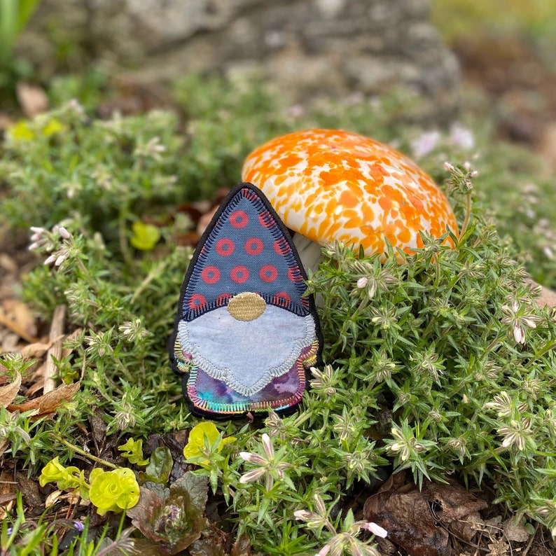 May include: A whimsical gnome patch with a blue hat adorned with red circles, a gray beard, and a colorful tunic. The patch is set against a backdrop of lush green foliage and a vibrant orange and white mushroom.