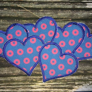 May include: A collection of heart-shaped fabric patches. Each patch features a blue background with a pattern of pink circles. The edges are outlined with a dark blue stitching. The patches are arranged on a weathered wooden surface.