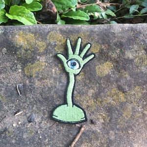 May include: A green embroidered patch featuring a hand with an eye in the palm. The patch has a long stem and a rounded base. The eye is blue with a black pupil. The patch is on a textured gray surface.