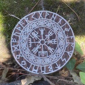 May include: A round, brown and white embroidered patch featuring a Viking compass design, also known as the Vegvísir. The patch has a circular border with runic symbols. The Vegvísir symbol is in the center, surrounded by intricate knotwork.
