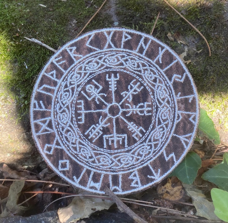 May include: A circular, brown and white embroidered patch with a Viking compass design, or Vegv&iacute;sir. The patch has a border of runic symbols. The Vegv&iacute;sir symbol is in the centre, surrounded by detailed knotwork.