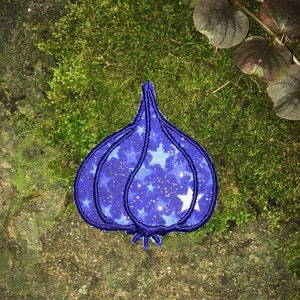 May include: A blue embroidered patch in the shape of a garlic clove. The patch features a starry night design with white stars on a dark blue background. The patch is on a mossy surface.