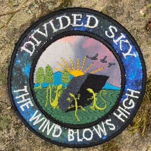 May include: Embroidered patch with the text "DIVIDED SKY THE WIND BLOWS HIGH." The patch features a landscape with a black geometric shape, yellow sun, green trees, and stylized lizards. The border has a starry night design.