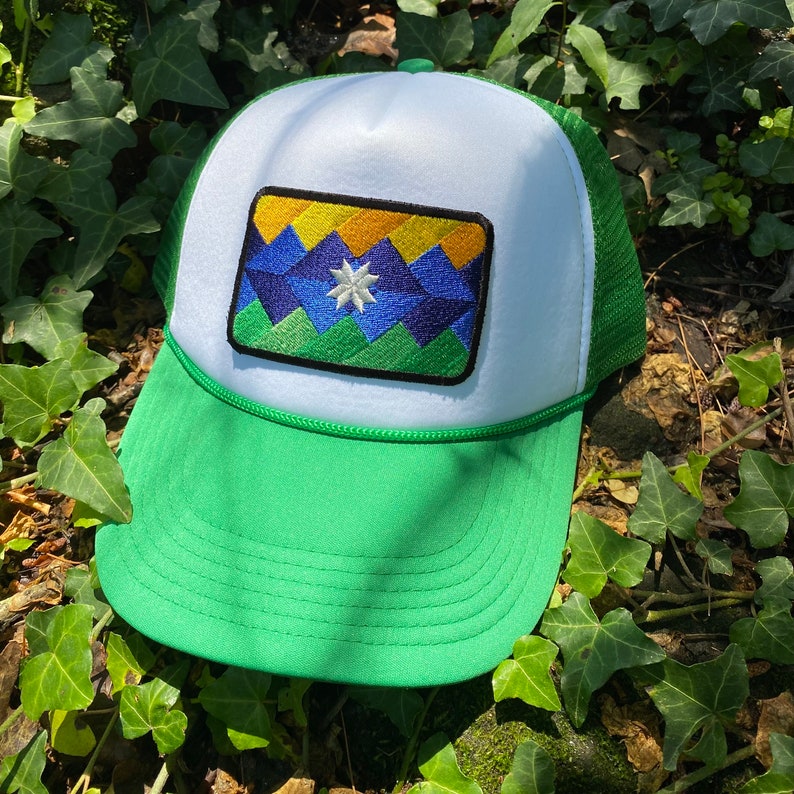 May include: A green and white trucker hat with a colorful embroidered patch. The patch features a geometric mountain design in shades of blue, green, yellow, and orange, with a white star in the center. The hat has a green brim and mesh back.