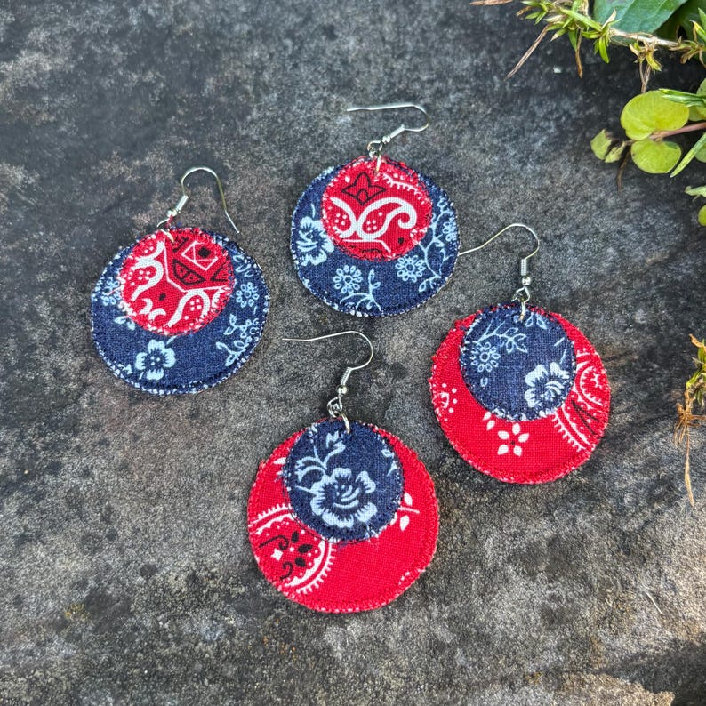 May include: Four pairs of round fabric earrings. Each earring features a red paisley pattern, a navy blue floral pattern, and silver-colored ear wires. The earrings are arranged on a textured, gray surface.