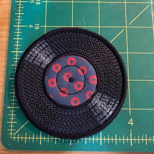 Pu&ograve; includere: Una toppa circolare con un anello esterno nero intrecciato e un cerchio centrale pi&ugrave; piccolo. Il cerchio centrale &egrave; blu navy con un motivo di cerchi rossi. La toppa &egrave; su un tappetino da taglio verde.