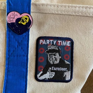 K&ouml;nnte beinhalten: Beige Canvas-Tragetasche mit blauem Riemen und zwei Aufn&auml;hern. Ein Aufn&auml;her ist herzf&ouml;rmig mit einem Totenkopf und den Worten "Live Laugh Love". Der andere Aufn&auml;her sagt "Party Time" und zeigt einen Mann, der einen Daumen hoch zeigt.