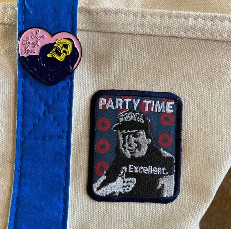 K&ouml;nnte beinhalten: Beige Canvas-Tragetasche mit blauem Riemen und zwei Aufn&auml;hern. Ein Aufn&auml;her ist herzf&ouml;rmig mit einem Totenkopf und den Worten "Live Laugh Love". Der andere Aufn&auml;her sagt "Party Time" und zeigt einen Mann, der einen Daumen hoch zeigt.