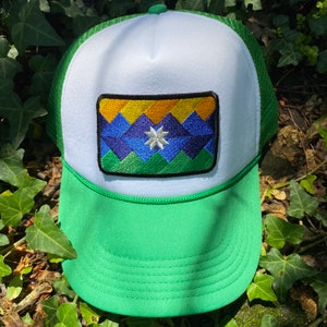 May include: A green and white trucker hat with a colorful embroidered patch. The patch features a mountain range design in shades of blue, yellow, and green, with a white star in the center. The hat has a green brim and mesh back.