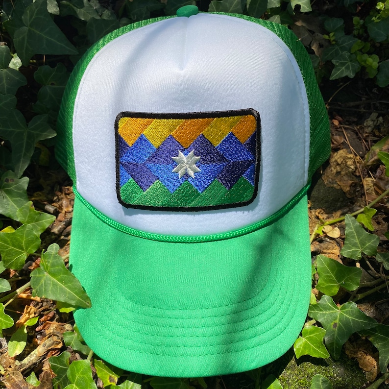 May include: A green and white trucker hat with a colorful embroidered patch. The patch features a mountain range design in shades of blue, yellow, and green, with a white star in the center. The hat has a green brim and mesh back.