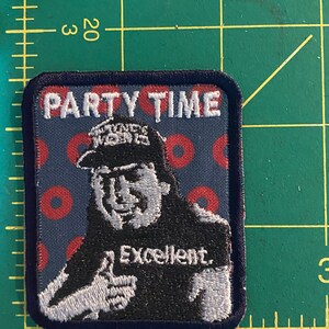 K&ouml;nnte beinhalten: Gestickter Aufn&auml;her mit dem Text "PARTY TIME" &uuml;ber einem Cartoon-Bild eines Mannes, der einen Daumen hochh&auml;lt. Der Mann tr&auml;gt einen Hut und ein T-Shirt mit dem Wort "Excellent."