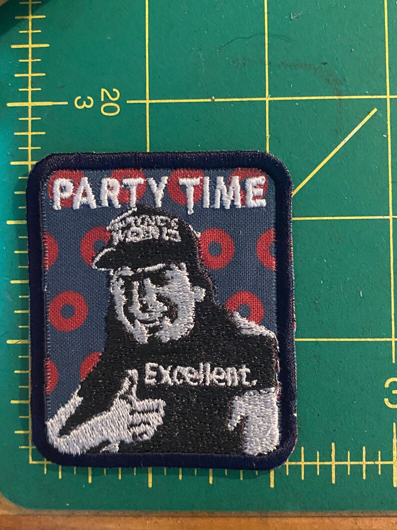 K&ouml;nnte beinhalten: Gestickter Aufn&auml;her mit dem Text "PARTY TIME" &uuml;ber einem Cartoon-Bild eines Mannes, der einen Daumen hochh&auml;lt. Der Mann tr&auml;gt einen Hut und ein T-Shirt mit dem Wort "Excellent."