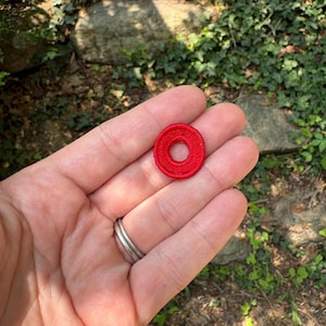 May include: A small, circular red embroidered patch featuring a central hole, creating a ring-like shape. The embroidery consists of dense, uniform red thread stitches covering the entire surface of the circular fabric piece.