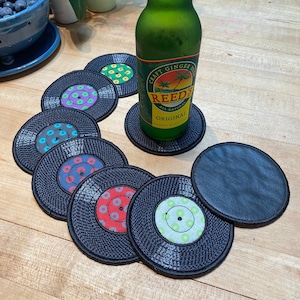Pu&ograve; includere: Un set di sottobicchieri neri a forma di dischi in vinile con etichette centrali colorate. Una bottiglia di vetro verde di Reed's All Natural Original ginger beer &egrave; appoggiata su un sottobicchiere. I sottobicchieri sono disposti su una superficie di legno.