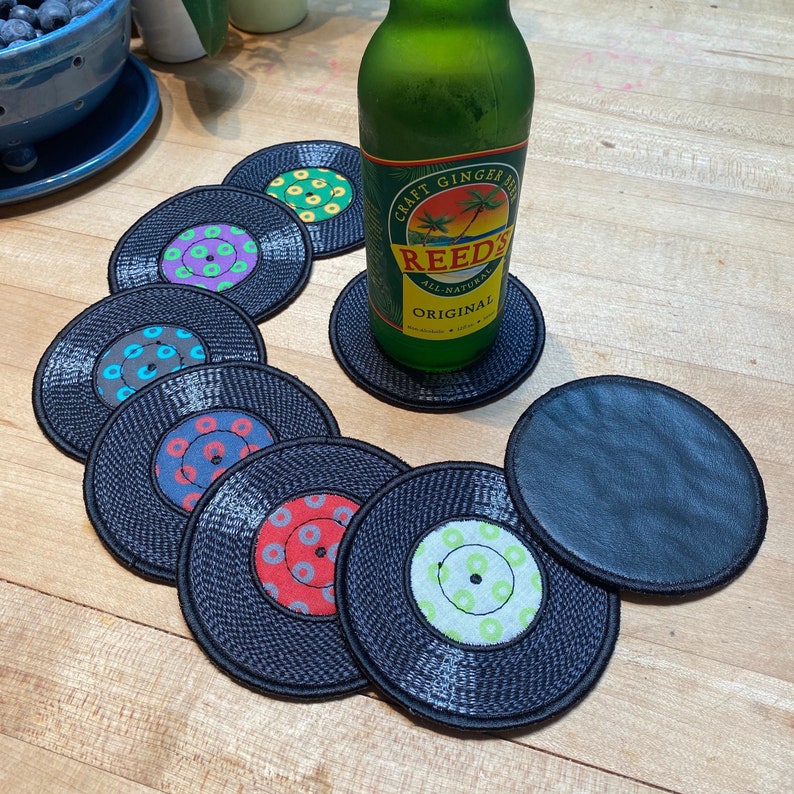 Pu&ograve; includere: Un set di sottobicchieri neri a forma di dischi in vinile con etichette centrali colorate. Una bottiglia di vetro verde di Reed's All Natural Original ginger beer &egrave; appoggiata su un sottobicchiere. I sottobicchieri sono disposti su una superficie di legno.