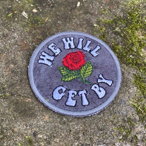 May include: Circular patch with the text "WE WILL GET BY" in light blue, surrounding a red rose with green leaves. The patch has a gray background and a gray border.