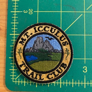 Op de afbeelding: Ronde geborduurde patch met de tekst "MT. ICCULUS TRAIL CLUB". De patch toont een landschap met bergen, een meer en een vogel tegen een blauwe lucht. De rand is beige met zwarte tekst en een zwarte omtrek.