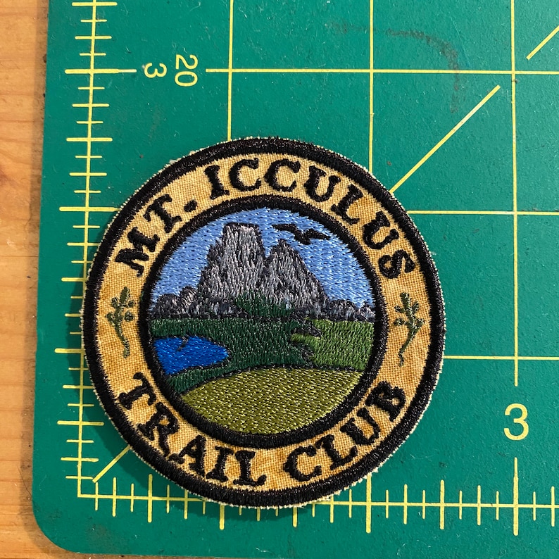 Op de afbeelding: Ronde geborduurde patch met de tekst "MT. ICCULUS TRAIL CLUB". De patch toont een landschap met bergen, een meer en een vogel tegen een blauwe lucht. De rand is beige met zwarte tekst en een zwarte omtrek.