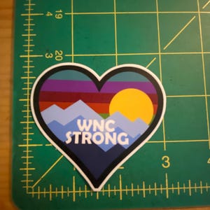 Puede incluir: Adhesivo en forma de coraz&oacute;n con un dise&ntilde;o de monta&ntilde;a y puesta de sol. El adhesivo presenta una cordillera azul, un sol amarillo y el texto "WNC STRONG" en blanco. El fondo tiene rayas horizontales de varios colores.