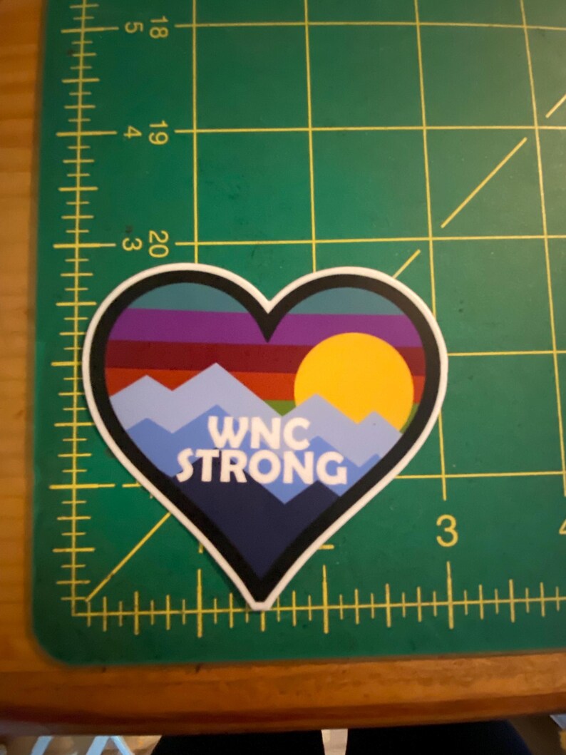 Puede incluir: Adhesivo en forma de coraz&oacute;n con un dise&ntilde;o de monta&ntilde;a y puesta de sol. El adhesivo presenta una cordillera azul, un sol amarillo y el texto "WNC STRONG" en blanco. El fondo tiene rayas horizontales de varios colores.