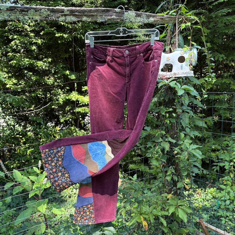 Op de afbeelding: Bordeauxkleurige corduroy broek met patchwork detail aan de zoom. Het patchwork heeft verschillende kleuren en patronen, waaronder bloemen en abstracte vormen. De broek hangt buiten aan een kledinghanger.